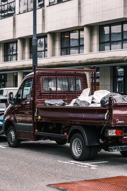 A small maroon flatbed truck parked on a city street, loaded with various types of discarded rubbish in black and grey plastic bags, along with loose debris. The truck has a metal mesh guard at the cab’s rear, which is slightly rusted, and the bed is filled to capacity. The vehicle is positioned in front of a modern multi-story building with large glass windows and concrete panels, reflecting the urban environment. The surrounding area features a pavement with a white painted marking, and nearby vehicles can be seen, including a white van parked further along the street. The scene appears to be during daytime with overcast lighting, and the detailed materials and textures of the rubbish bags highlight the contrast between the commercial environment and the waste materials prepared for collection, illustrating a typical scene related to private rubbish removal or on-site clearance services offered by companies like rubbishclearancekingstonuponthames.co.uk amidst a busy city setting.