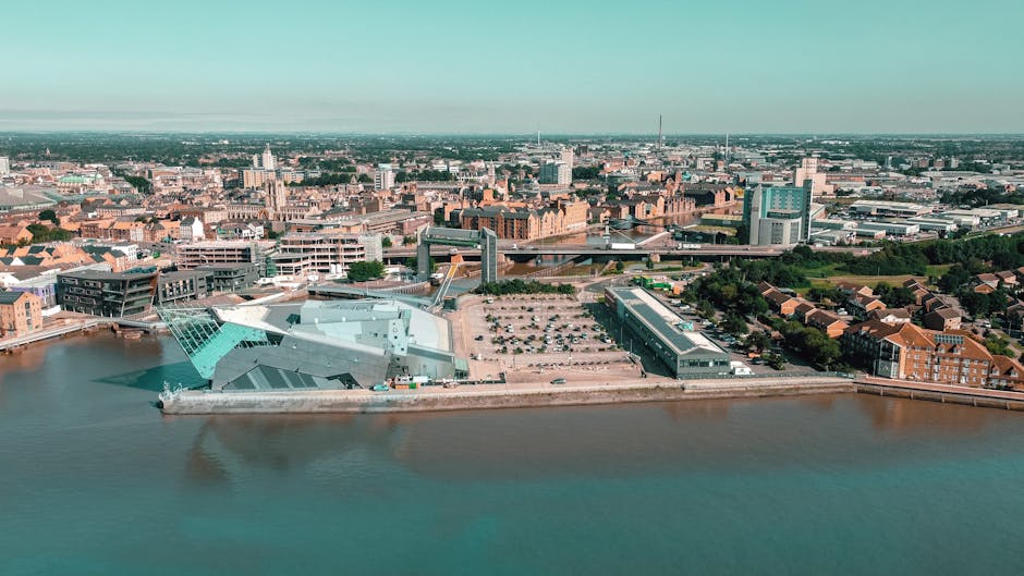 An aerial view of a cityscape showing modern and historic buildings near a wide river. In the foreground, there is a large, angular structure with a metallic, reflective surface, possibly a contemporary architectural building used for events or exhibitions. Adjacent to it, a parking lot with several vehicles is visible, situated next to a long, low-rise building with a dark roof and glass windows, likely a commercial or storage facility. Surrounding the area are densely packed residential houses with red brick facades and tiled roofs, characteristic of the local architecture. In the background, the skyline is populated with mid-rise office buildings, some with glass exteriors, and prominent church spires and towers, reflecting the urban environment. The scene is illuminated by daylight, with a clear sky, conveying a calm, neutral atmosphere. The image subtly emphasizes the importance of proper waste management and disposal, consistent with independent rubbish removal services like those offered in Kingston upon Thames, and showcases the integration of modern infrastructure within the city environment.