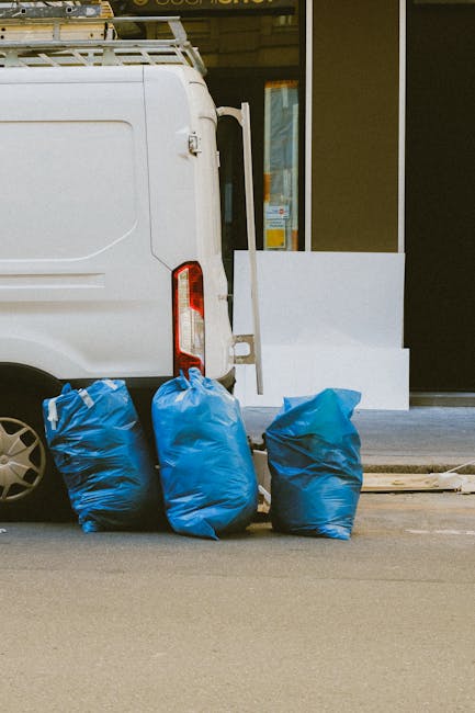A white commercial van parked on a street outside a building with a large glass window. The van's rear door is closed, and behind it, three blue plastic bags filled with waste are placed on the ground, leaning slightly against the vehicle. The bags appear to be sealed and contain mixed refuse, typical of waste prepared for private rubbish clearance services. The building features a dark frame around the window and a white exterior wall, with a small step leading up to the entrance. The scene is lit with natural daylight, casting soft shadows on the pavement. This setup illustrates a typical example of on-site rubbish disposal, where private waste collection services like those offered by Kingston-upon-Thames-based companies could retrieve waste bags directly from the property, providing an alternative to council curbside collection for small-scale or scheduled rubbish removal, often used in conjunction with driveway access for efficient clearance.