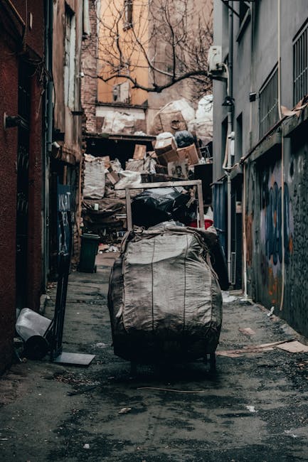 A narrow urban alleyway cluttered with piled rubbish and discarded debris, including cardboard boxes, black plastic bags, and miscellaneous waste materials, set against the backdrop of brick and concrete buildings on either side. In the foreground, a large, grey, weathered wheeled rubbish bag is positioned in the middle of the alley, with its handles and fabric showing signs of use. The ground is uneven, stained, and strewn with small pieces of trash. Behind the pile, there are uneven stacks of accumulated waste, possibly awaiting collection or disposal, with some boxes leaning against the clutter. The alley is partially enclosed by high walls, some decorated with graffiti and peeling paint, with bare tree branches visible overhead, suggesting an outdoor setting. Ambient lighting indicates overcast weather, emphasizing the gritty and neglected atmosphere typical of areas requiring rubbish removal services, such as those managed by companies like rubbishclearancekingstonuponthames.co.uk, which provide alternative waste handling solutions outside regular municipal collection.