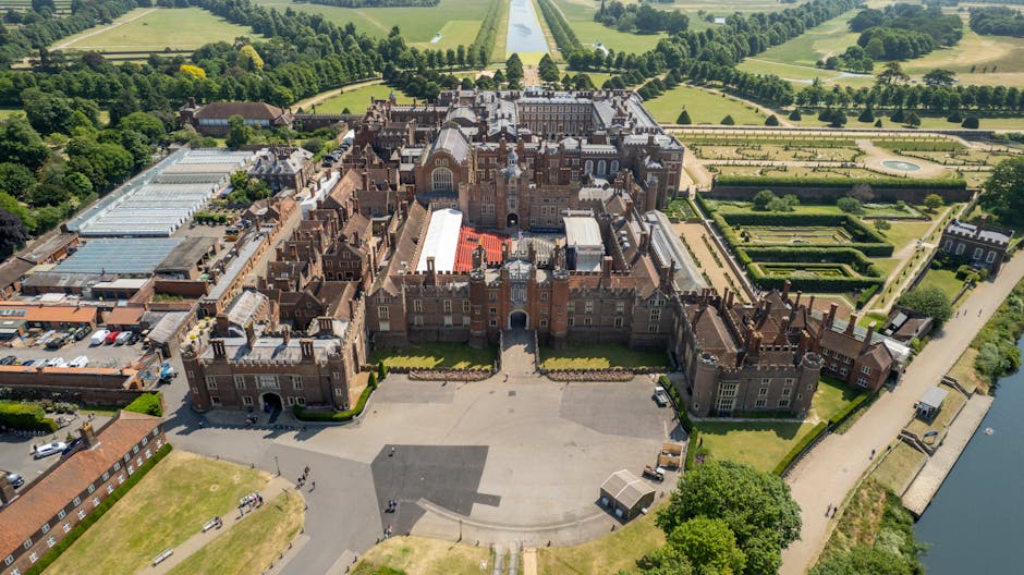 An aerial view of Hampton Court Palace showing its historic red brick and stone façade with numerous chimneys, turrets, and gabled roofs. The palace's expansive courtyards are visible, with the main entrance front and center. Surrounding the palace are neatly manicured gardens, tree-lined pathways, and formal lawns extending into the distance. To the left of the palace, there are several modern structures, possibly service or maintenance buildings, with flat roofs and a mix of brick and metal finishes. In front of the palace, a large paved courtyard area slopes downward, with a small, sheltered structure on one side. Nearby, there are visible parking areas and a few vehicles. The environment is lush, with dense green trees framing the historic site and open fields further beyond. The scene is well-lit, suggesting clear weather, with natural sunlight casting shadows across the landscape. This aerial perspective highlights the contrast between the historic architecture and the surrounding landscaped gardens, typical of a site that might receive private or independent waste management services for event logistics or waste clearance related to site maintenance, supported by companies specializing in rubbish removal.