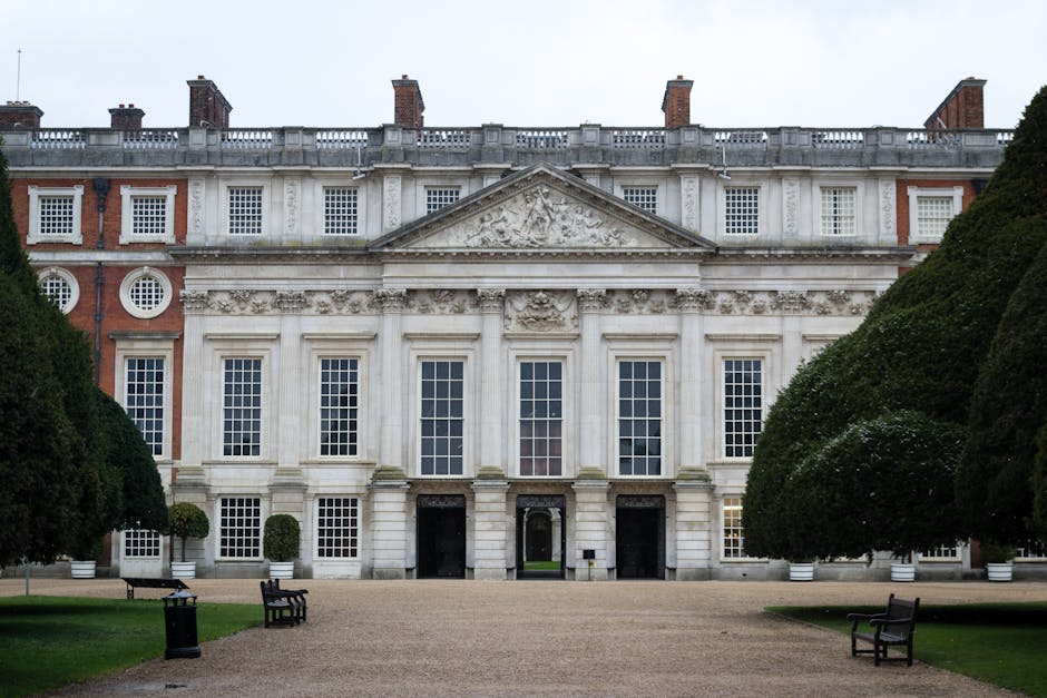 An aerial view of Hampton Court Palace showing its historic red brick and stone façade with numerous chimneys, turrets, and gabled roofs. The palace's expansive courtyards are visible, with the main entrance front and center. Surrounding the palace are neatly manicured gardens, tree-lined pathways, and formal lawns extending into the distance. To the left of the palace, there are several modern structures, possibly service or maintenance buildings, with flat roofs and a mix of brick and metal finishes. In front of the palace, a large paved courtyard area slopes downward, with a small, sheltered structure on one side. Nearby, there are visible parking areas and a few vehicles. The environment is lush, with dense green trees framing the historic site and open fields further beyond. The scene is well-lit, suggesting clear weather, with natural sunlight casting shadows across the landscape. This aerial perspective highlights the contrast between the historic architecture and the surrounding landscaped gardens, typical of a site that might receive private or independent waste management services for event logistics or waste clearance related to site maintenance, supported by companies specializing in rubbish removal.
