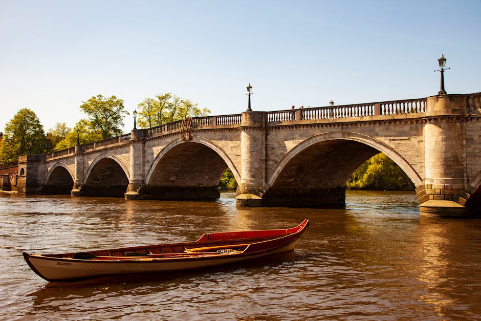 A red and white rowing boat floats in calm water beneath a stone bridge with multiple arches, which features decorative lamp posts along its balustrade. The bridge's stone surface has a weathered, textured appearance, and the water below reflects the bridge's arches and the boat. In the background, there are lush green trees with leaves in varying shades of green, indicating a spring or summer setting. The sky above is clear with a light blue tone, and the scene is illuminated by natural daylight, casting soft shadows. The setting appears to be a serene river location, possibly used for leisure activities or private boat storage, with an environment suitable for independent or on-site waterborne transportation. The overall scene emphasizes a peaceful riverside environment with a historic stone bridge, aligning with a context of local or private watercraft management and scenic outdoor spaces.