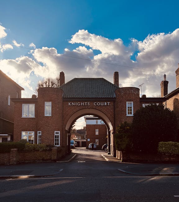 A daytime view of a brick residential complex named Knights Court with an arched entranceway, situated on a paved street. The arch is constructed of red bricks and features the name 'KNIGHTS COURT' in white letters at its top. Flanking the arch are two small towers with rounded tops, also made of red brick. Behind the arch, there are parked cars visible in the background, along with additional brick buildings, some with chimneys. The sky above displays a mix of blue and white, with large, fluffy clouds illuminated by sunlight creating a contrast against the darker shaded areas of the buildings and trees. On either side of the street, low brick walls and neatly trimmed bushes frame the scene, complementing the residential environment. This image visually relates to private property and on-site clearance contexts often associated with independent rubbish removal services like those offered by rubbishclearancekingstonuponthames.co.uk, focusing on the outdoor setting typical of rubbish collection or disposal operations.