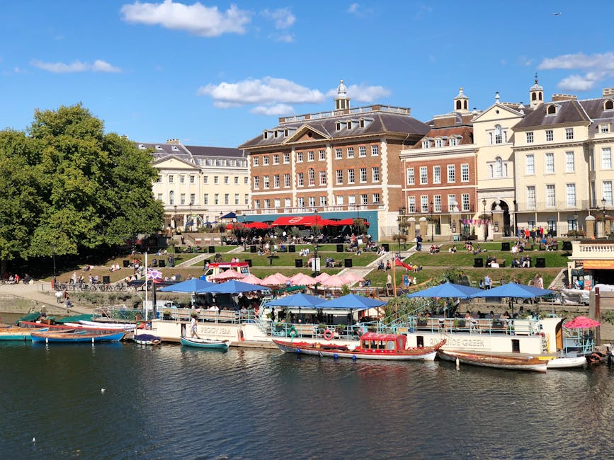 A riverside scene featuring multiple small boats moored along the edge of the water, including a larger vessel with a white hull and a red and white striped canopy, and smaller boats with blue canopies and wooden finishes. Behind the boats, there is a busy outdoor café area with pink and blue umbrellas providing shade for patrons seated at tables on a terraced grassy slope. Several people are visible walking along pathways or sitting on benches, overlooking the water. To the left, a large green leafy tree partially obscures a series of multistory historic buildings with classical architectural details, including brick and cream-colored facades, decorative cornices, and dome-style rooftops. The scene is illuminated by bright daylight with a clear blue sky and scattered white clouds, creating a lively and scenic environment that might relate to independent or leisure activities near a waterfront, similar to alternative waste management or private clearance services such as those provided by Kingston upon Thames rubbish clearance companies.