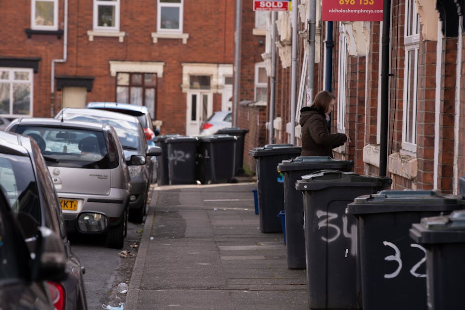 A residential street scene in a suburban area with brick terraced houses featuring white window frames and stone window sills. Several black wheelie bins lined up along the narrow sidewalk are visible, some with lids partially open or closed, and they are marked with white spray-painted numbers. A woman dressed in a dark brown hooded coat is standing beside one of the bins, appearing to be in the process of disposing of rubbish. The street is lined with parked cars, including a silver hatchback with a visible UK registration plate on the left side of the image. In the background, additional houses and more wheelie bins can be seen, with some bins placed directly in front of the properties. The scene is illuminated by natural daylight, with an overcast sky creating diffuse lighting. The image depicts a practical scene of on-site rubbish disposal, possibly related to informal waste collection or private clearance, as suggested by the arrangement of the bins outside the residences.