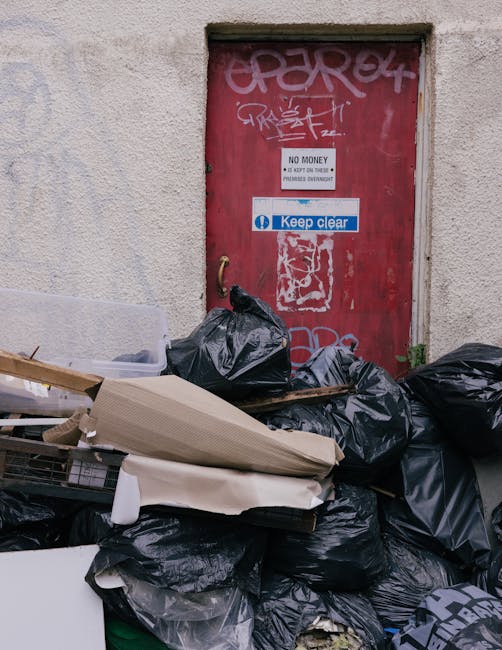 A large pile of black garbage bags, some torn or partially open, is piled up in front of a red metal door set into a beige textured wall. The waste bags contain a mixture of household items and discarded materials. Among the debris, there are pieces of cardboard, a canvas or fabric item, and parts of wooden furniture or construction rubbish. The door features multiple layers of graffiti markings in white and pink spray paint, along with a white sign indicating 'No Money' and a smaller blue and white sticker that reads 'Keep clear.' The scene appears to be an outdoor area, possibly a back alley or service entrance, where refuse has been temporarily dumped, consistent with private or independent rubbish disposal practices that some residents or waste management services like rubbishclearancekingstonuponthames.co.uk might handle as an alternative to official council rubbish collection. The general atmosphere indicates an unorganized waste accumulation, highlighting the importance of proper rubbish clearance and disposal regulations.