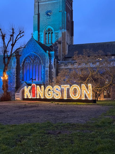 A nighttime view of a historic brick church with tall, pointed Gothic-style windows illuminated in blue light, situated behind a large, illuminated sign spelling out 'KINGSTON' in colorful, glowing letters. The sign is positioned on a grassy area with some bare trees nearby, indicating a winter setting. The scene appears outside an urban or town center environment, with the church serving as a notable architectural feature, and the lighting emphasizing its structural details. This setting relates to local community identity and could be associated with nearby public spaces or cultural landmarks, reflecting how non-official approaches to waste disposal might be considered alongside traditional methods. The overall scene offers a calm, dusk atmosphere, with subtle artificial lighting highlighting the church and signage in an area that might be used for public events or local visits, subtly connecting the environment to the context of private waste handling and alternative rubbish collection options.