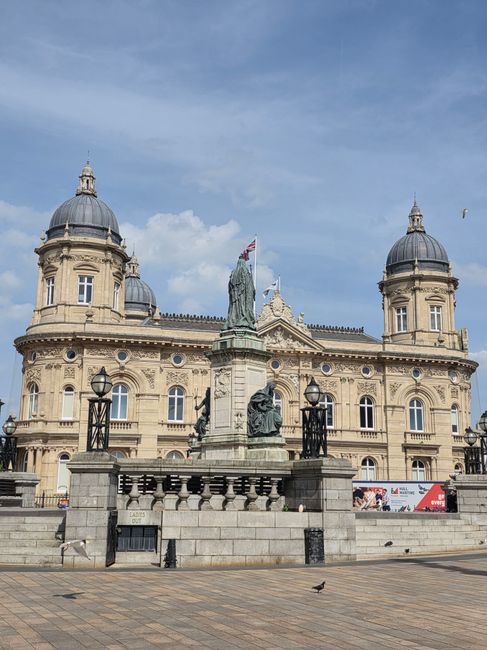 A historic sandstone building with symmetrical architectural features, including two prominent domed towers on either side, and ornate decorative elements along the roofline, situated behind a paved terrace. In front of the building, a stone monument features an equestrian statue and a seated figure, flanked by black wrought iron lamp posts. The scene is illuminated by natural daylight with a clear blue sky and a few scattered clouds in the background; a single pigeon is visible on the pavement. The environment appears to be an open public space or square, with minimal surrounding structures visible. Given the context of rubbish removal services, this image may evoke considerations of maintaining cleanliness around historic sites or public landmarks, highlighting the importance of proper waste disposal during urban and cultural site management, as handled by providers like [COMPANY_NAME]. The scene emphasizes the importance of on-site clearance and private waste handling in sensitive or high-profile locations, aligning with the theme of alternative waste management options versus municipal rubbish collection.