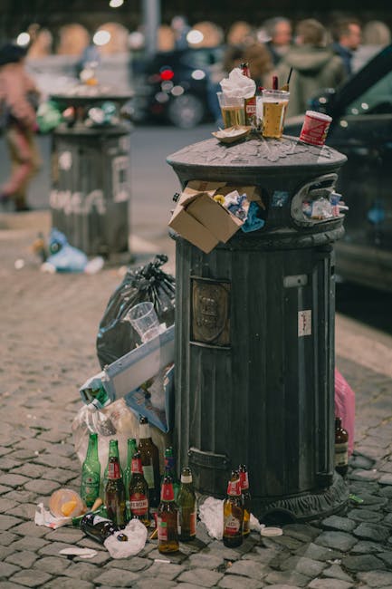 A black, cylindrical public litter bin made of metal with a decorative domed top, situated on a cobblestone street. The bin is overflowing with various waste items, including crumpled paper, cardboard, and plastic bottles, some of which are on the ground around it. The top of the bin holds several discarded coffee cups with plastic lids and straws, a paper food container, and a small red can, indicating recent use. A black plastic trash bag leans against the side of the bin, and additional bottles, including glass beer bottles and plastic soft drink bottles, are scattered nearby, some with lids or caps still attached. In the background, there are blurred figures of people, parked cars, and a grey metal waste container, suggesting an active street scene. The environment appears to be urban with foot traffic, and the visible litter relates to typical rubbish accumulated from social gatherings or street-side consumption, highlighting the importance of rubbish removal services like those offered by Rubbish Clearance Kingston upon Thames in providing alternative waste handling solutions outside of municipal collection systems.