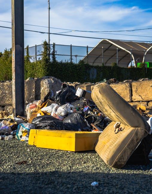 A large outdoor collection of bulk waste and cardboard packaging, primarily consisting of stacked boxes of fresh fruit, with some black plastic crates on top, situated next to various recycling and general waste bins in green, red, and grey colours. The boxes of fruit packaging are made of cardboard with colorful printed labels, and are arranged in multiple stacks, some leaning slightly. To the right, there are metal wire cages filled with miscellaneous items, including brown cardboard boxes and smaller packaged goods, indicating a collection area likely for private rubbish removal or on-site clearance. The background shows a rural environment with trees and a house visible under a bright sky, with natural daylight illuminating the scene and creating clear, distinct shadows. This scene reflects typical arrangements used by independent waste clearance services, such as those offered by companies like Rubbish Clearance Kingston upon Thames, and highlights a mixture of packaging waste and other recyclable materials awaiting disposal.