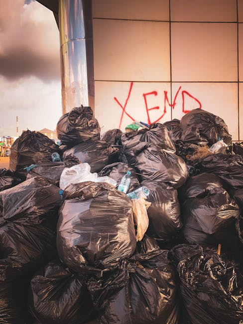 A large pile of tightly tied black rubbish bags occupying most of the foreground and midground, placed against a modern tiled wall with light-colored rectangular tiles. Some bags appear bulging with waste, and a few transparent plastic water bottles are visible among the bags, indicating mixed household or general waste. In the background, part of a cityscape with low-rise buildings and a cloudy sky is visible, suggesting an urban environment. There is red graffiti on the tiled wall with the text 'XEND,' adding an element of street art to the scene. The scene is outdoors, likely in a service area or back alley, with natural daylight illuminating the scene softly, emphasizing the textures of the plastic bags and the reflective surface of the tiled wall. This image captures an example of waste collection or disposal, relevant to independent rubbish removal services often used for on-site clearance or alternative waste handling in urban settings, as provided by companies like rubbishclearancekingstonuponthames.co.uk.
