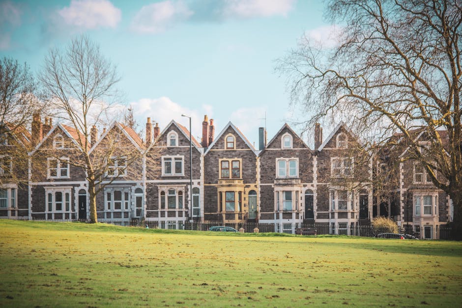 A row of traditional Victorian-style terraced houses constructed from grey and brown brickwork, featuring steep gabled roofs with slate tiles, white-framed sash windows, and decorative stone detailing around doorways and windows. The houses are set along a quiet street with a front garden or yard area, and a low wooden fence runs parallel to the pavement. In the foreground, there is a well-maintained grassy field with scattered fallen leaves, and a few leafless trees with bare branches stretching upward, indicating a late autumn or winter setting. The sky above is light blue with some scattered clouds, and ambient daylight provides a clear, neutral atmosphere. The scene, captured outdoors in daylight, suggests a peaceful residential environment where local authority or private rubbish removal services such as those provided by Kingston-based waste management companies may be involved in house clearance or rubbish collection activities nearby.